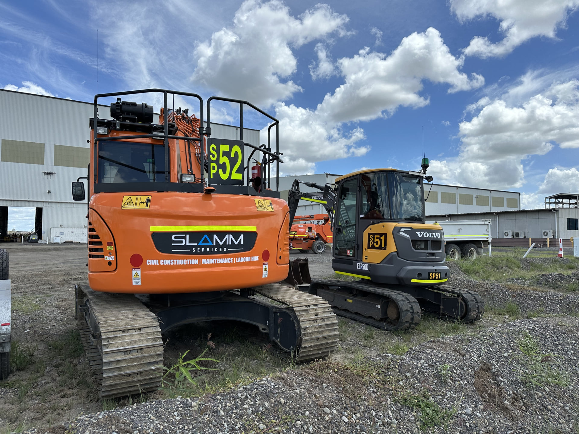An orange Slamm Services excavator (SP52) and a yellow Volvo ECR50D excavator (SP51) parked on a gravel lot near an industrial building under a partly cloudy sky.
