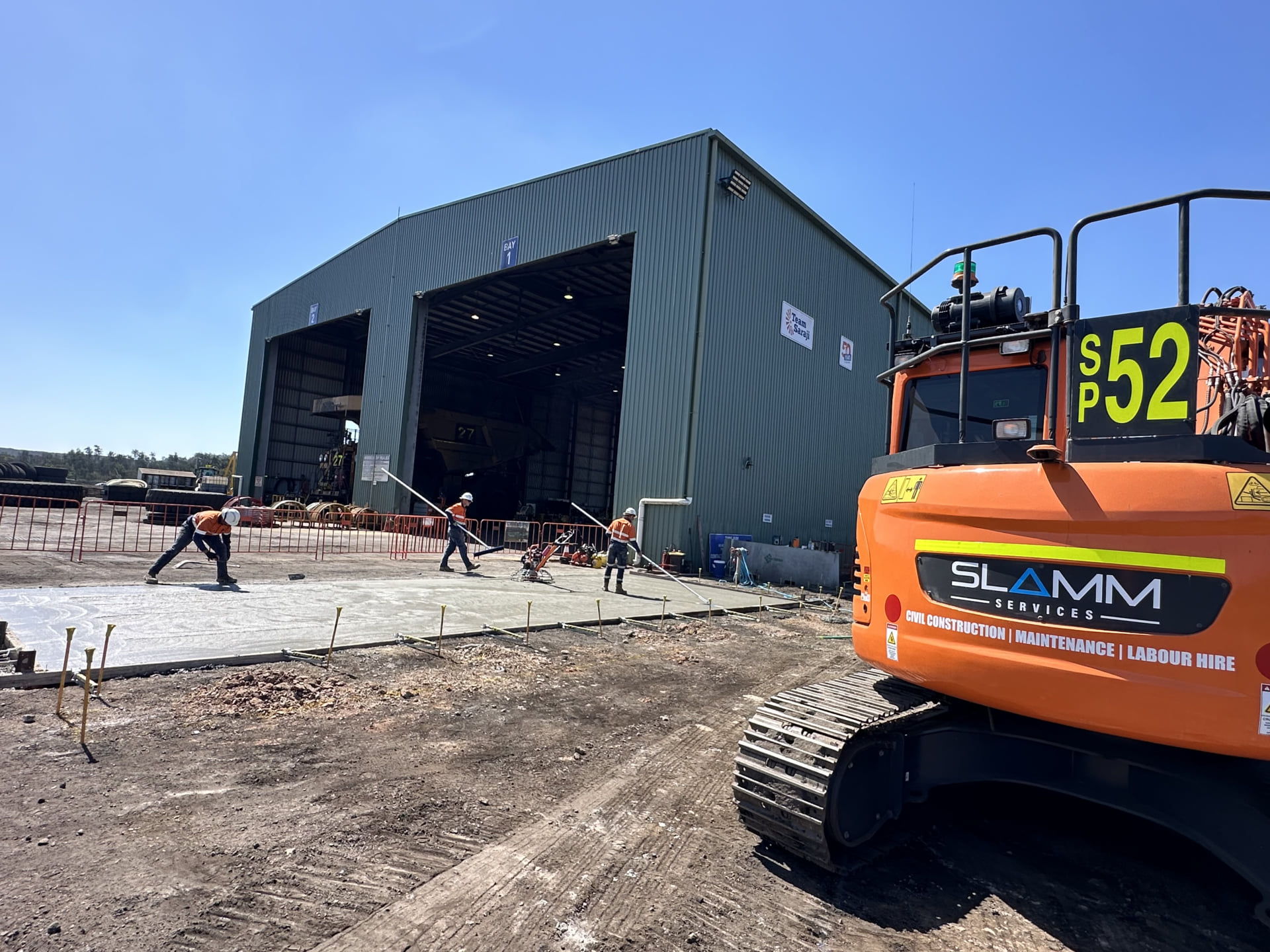 An orange Slamm Services excavator (SP52) and a yellow Volvo ECR50D excavator (SP51) parked on a gravel lot near an industrial building under a partly cloudy sky.